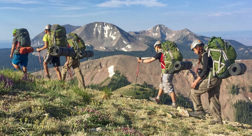 A group of students wearing backpacks hike along a trail. They appear to be at high elevation. There are mountains in the background. 